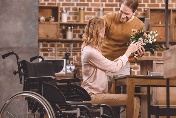 smiling man presenting flowers to disabled wife at home  - Foto, afbeelding