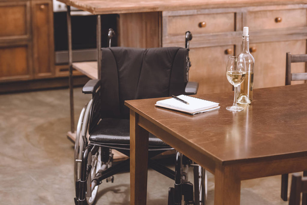 cadeira de rodas em pé perto da mesa com garrafa de vinho, óculos, caderno e caneta
 - Foto, Imagem