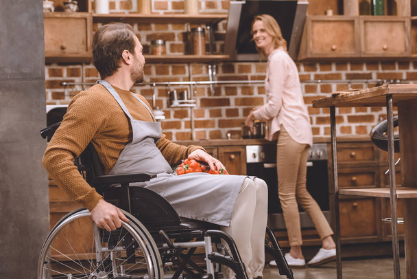 mujer sonriente mirando marido discapacitado en silla de ruedas sosteniendo verduras mientras cocinan la cena juntos en casa
   - Foto, imagen