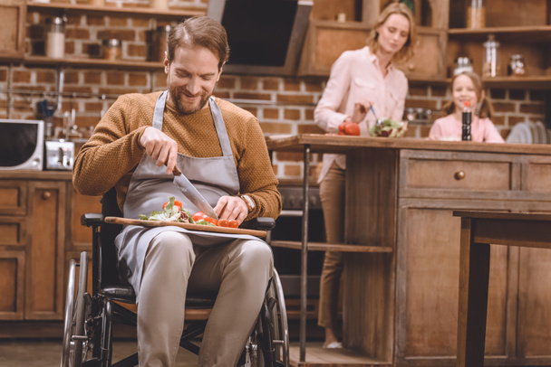 улыбающийся мужчина в инвалидной коляске режет овощи для салата, в то время как счастливые мать и дочь стоят позади
 - Фото, изображение