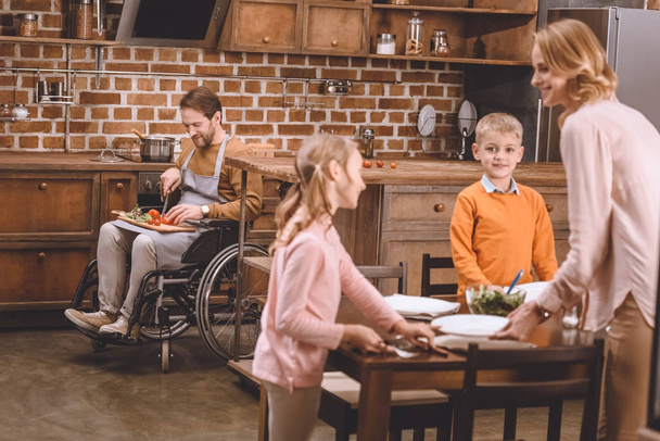 cute little kids with mother serving table for dinner while disabled father in wheelchair cutting vegetables behind   - Foto, Bild