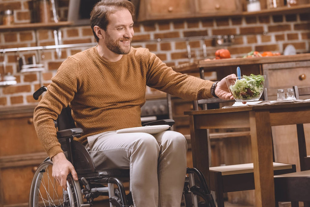 homme handicapé souriant en fauteuil roulant tenant bol en verre de salade de légumes à la maison
  - Photo, image