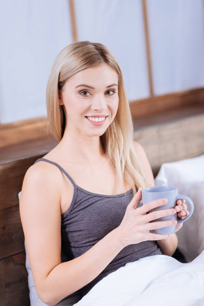 Cheerful young woman drinking morning coffee in bed - 写真・画像