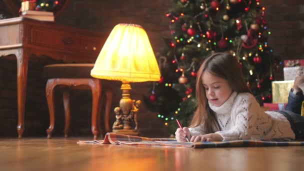 beautiful little girl in a white knitted sweater lies on the floor and writes a letter to Santa Claus, in the background a festive Christmas tree and a lot of presents - Кадри, відео