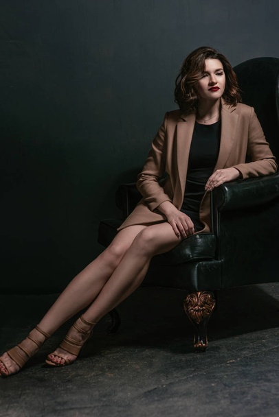 retrato de joven, hermosa mujer triste con pelo corto marrón con elegante maquillaje en vestido negro y chaqueta beige en sillón sobre fondo oscuro
 - Foto, imagen