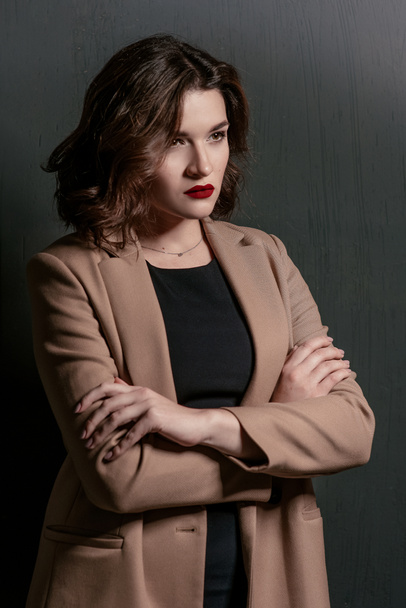portrait of young, beautiful sad woman with short brown hair with stylish make up in black dress and beige jacket on dark background - Foto, afbeelding