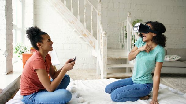 Mixed raced woman using virtual reality glasses while her friend holding digital tablet computer. Girlfriends play video game 3d technology concept at home - Foto, imagen