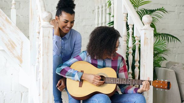 Two brazilian curly girls sistres sitting on stairs and practice to play acoustic guitar. Friends have fun and singing at home - Valokuva, kuva