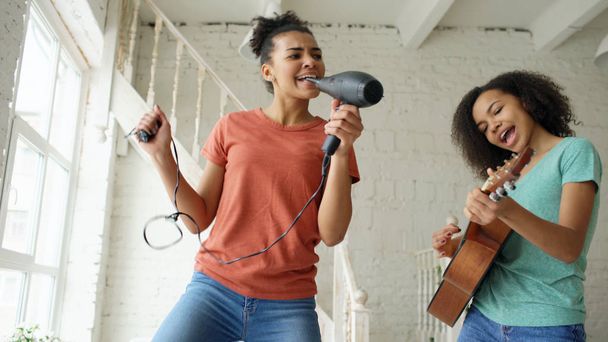 Mixed race young funny girls dance singing with hairdryer and playing acoustic guitar on a bed. Sisters having fun leisure in bedroom at home - Foto, afbeelding