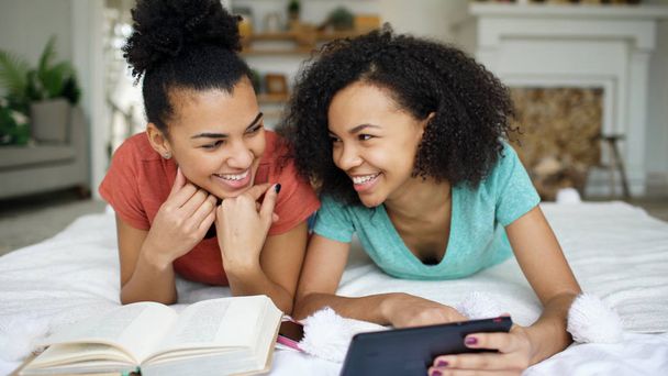 Two cheerful mixed race funny girlfriends reading book and using tablet computer talks and have fun lying in bed at home - Photo, Image