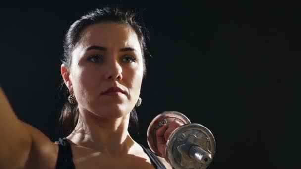 Young woman stands and raises metallic dumbbells up in her hands in a studio                      An encouraging view of a fitness instructor who stands and raises dumbbells in her hands up in an active and professional way in a black studio. - Filmagem, Vídeo