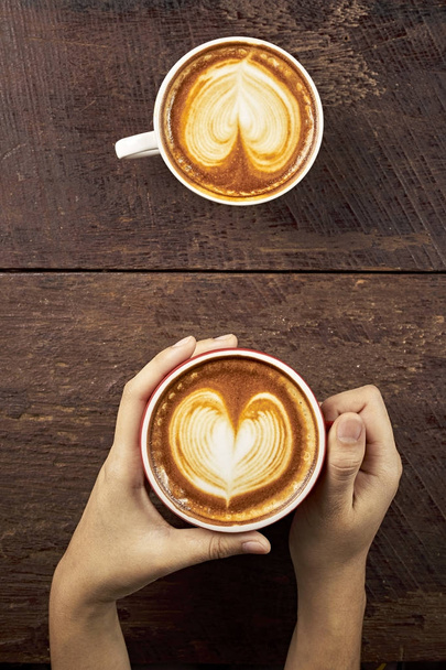 Руки женщины держат чашку кофе с пеной над деревянным столом, вид сверху - Фото, изображение