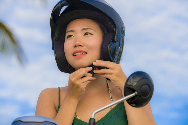 若い可愛い安全コンセプトの青い空に分離されたスクーター バイクに乗ってヘルメットを調整する幸せな、キュートなアジア中国女性 - 写真・画像