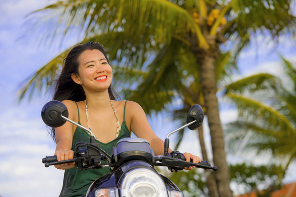 happy and pretty Asian Chinese woman having fun riding scooter  motorbike on tropical Summer palm tree background smiling cheerful  - Photo, Image
