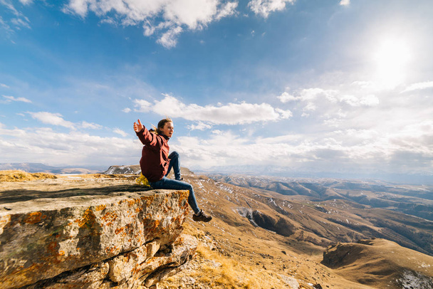 active young girl travels through the Caucasus mountains, sits on the edge of the cliff - Foto, Imagen
