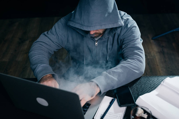mysterious man in a hood doing something illegal on a laptop, smoking - Foto, Imagem