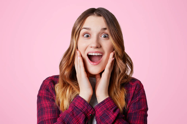 Photo of excited pretty woman looks in amazement, bugged eyes and widely opened eyes, being pleasantly shocked to recieve present, isolated over pink backgroud. Emotional female poses indoor - Photo, Image