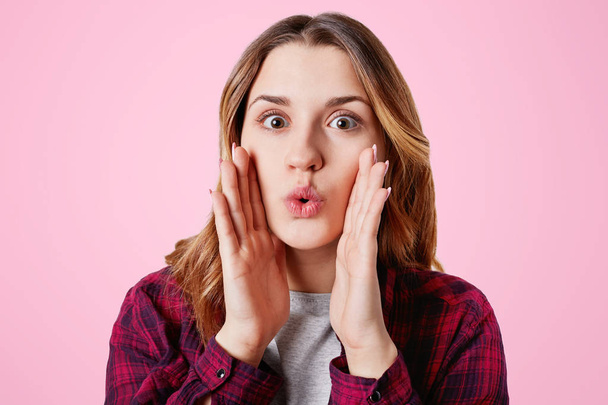 Amazed pretty lovely woman rounds lips as says wow, keeps hands on cheeks, being shocked, gestures in studio, isolated over pink background. Attractive young female model gossips with friends - Foto, Imagen