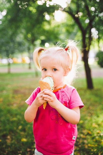little funny girl blonde eating sweet blue ice cream in a waffle cup on a green summer background in the park. smeared her face and cheeks and laughs. Dressed in bright stylish clothes - 写真・画像