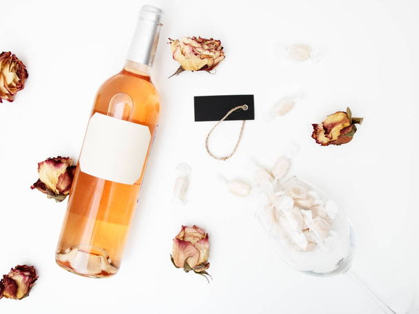 Επίπεδη θέσει. Μίνιμαλ στυλ. Μινιμαλιστική τάση φωτογραφίας. Ένα μπουκάλι ροζέ με μια σύνθεση από αποξηραμένα τριαντάφυλλα. Μπουκάλι κρασί, γυαλί και λευκό τραπέζι. Το Top view - Φωτογραφία, εικόνα