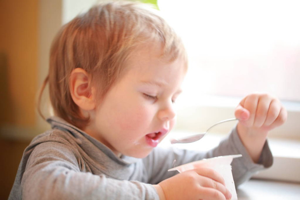 el niño come yogur con una cuchara sentada a la mesa
 - Foto, Imagen