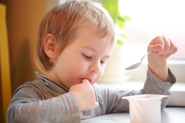 l'enfant mange du yaourt avec une cuillère assise à la table
 - Photo, image