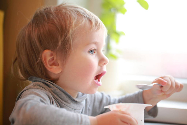 子供はテーブルに座ってスプーンでヨーグルトを食べる - 写真・画像