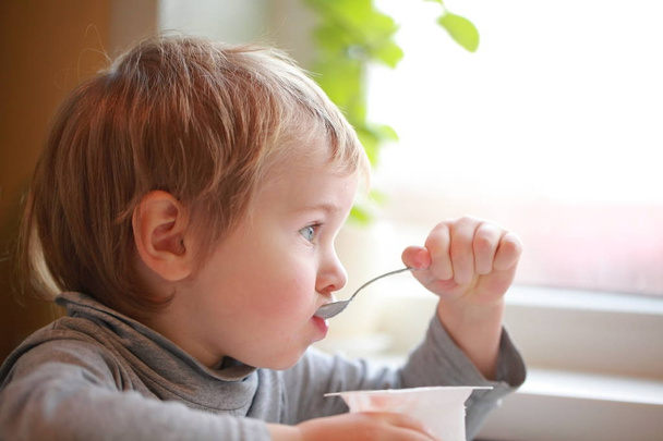 子供はテーブルに座ってスプーンでヨーグルトを食べる - 写真・画像