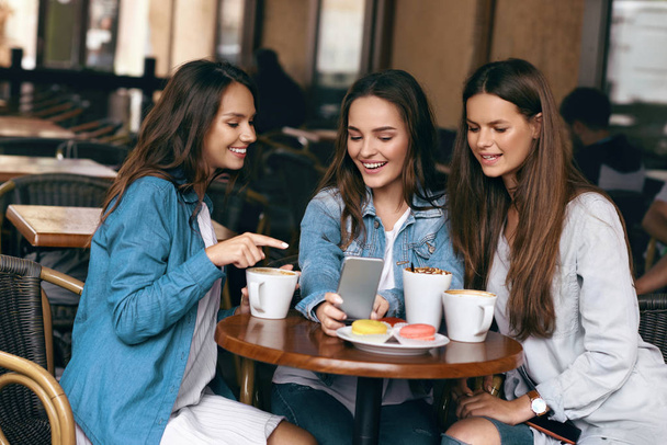 Young Female Friends Using Phone And Drinking Coffee In Cafe. - Foto, Bild