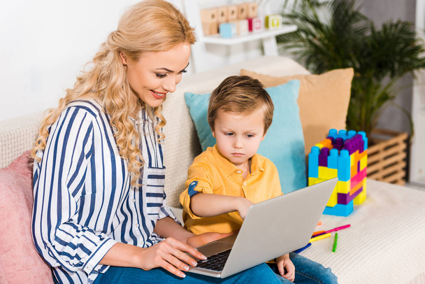 mosolyog, anya és kis fia együttes használata a laptop otthon a kanapén - Fotó, kép