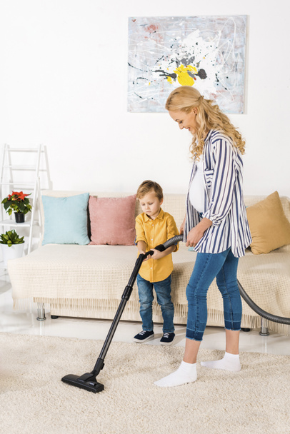 smiling pregnant woman with adorable little son cleaning carpet with vacuum cleaner together - Valokuva, kuva