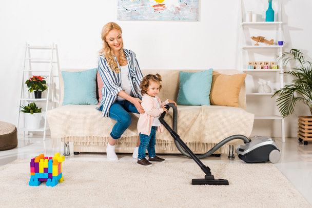 smiling pregnant woman looking at cute little daughter holding vacuum cleaner at home - Фото, зображення