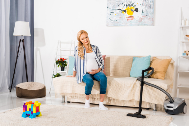 blonde pregnant woman sitting on sofa and looking at carpet with colorful blocks and vacuum cleaner - Photo, image