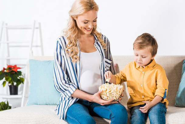 Schwangere und süßer kleiner Sohn essen zu Hause Popcorn aus Glasschüssel - Foto, Bild