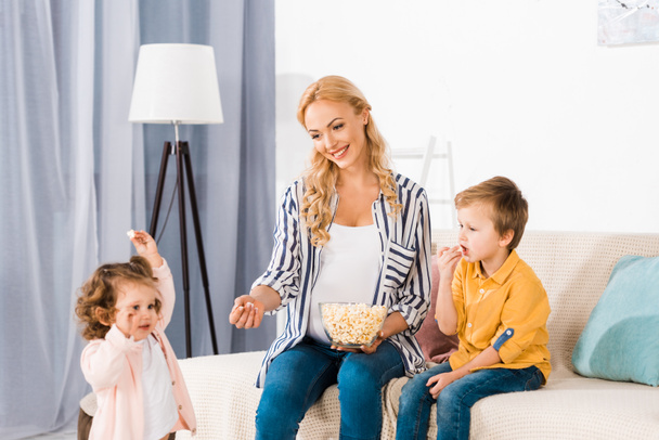 Gelukkig zwangere moeder en schattige kleine kinderen eten popcorn uit glazen kom thuis - Foto, afbeelding