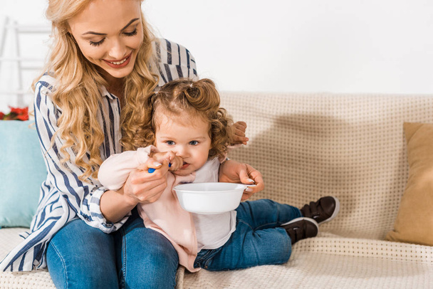 happy mother feeding adorable little daughter and holding plate with spoon at home - Photo, Image