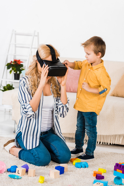 Netter kleiner Sohn schaut Mutter mit Virtual-Reality-Headset zu Hause an - Foto, Bild