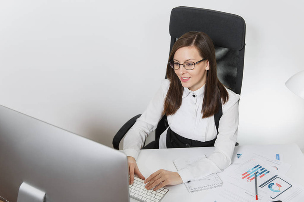 Mooie lachende brown-haar zakenvrouw in pak en bril zitten aan de balie, werken op computer met hedendaagse monitor met documenten in lichte office, handen op toetsenbord op witte achtergrond - Foto, afbeelding