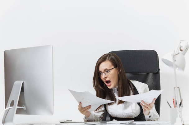 Pretty angry business woman in suit sitting at the desk with documents, working at computer with modern monitor in light office, swearing and screaming, resolving issues, on white background - Photo, Image