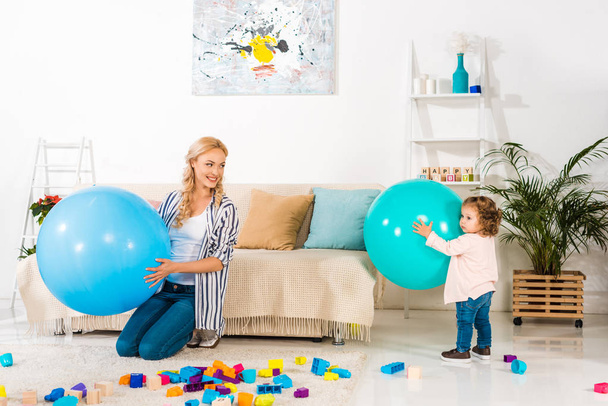 beautiful happy mother and infant daughter playing with fitness balls and colorful blocks at home - Фото, изображение
