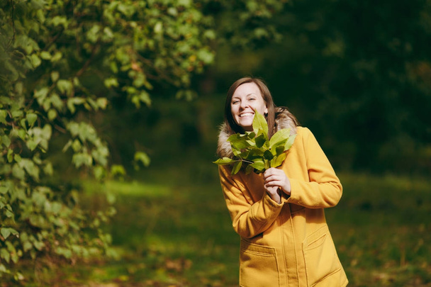 Mooie gelukkig Kaukasische lachende brown-haar jongedame in gele jas, jeans, laarzen in de groene bossen. Vrouwelijk model met gouden herfst bladeren staan en wandelen in de vroege herfst park buiten mode. - Foto, afbeelding