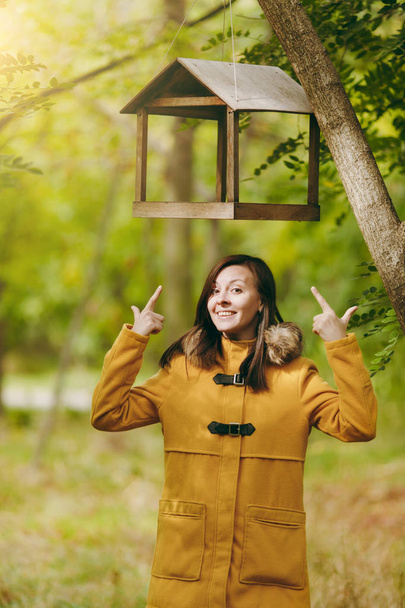 Szép boldog kaukázusi fiatal mosolygós barna hajú nő sárga kabát, farmer, bakancs, zöld erdőben. Őszi divat modell hagy madár etetők alatt áll a kora őszi park szabadban. - Fotó, kép