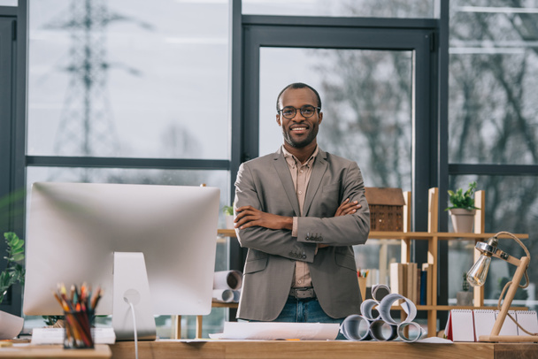 smiling african american architect with crossed arms standing at workplace with computer and blueprints - Фото, изображение