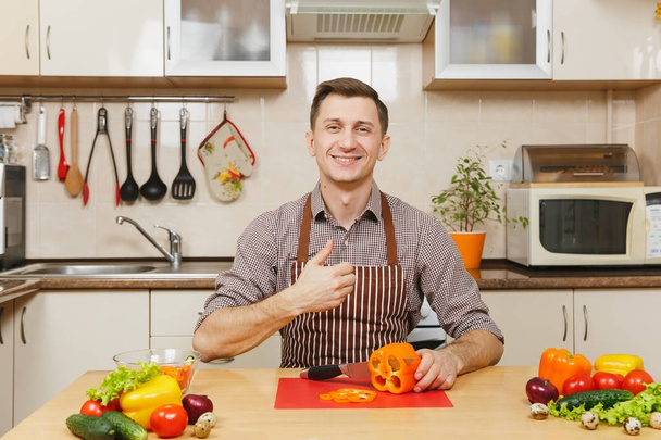 Guapo joven caucásico sonriente en un delantal, camisa marrón sentado a la mesa, mostrando el pulgar hacia arriba, corta verduras para ensalada con cuchillo en la cocina ligera. Concepto de dieta. Cocinar en casa. Preparar alimentos
. - Foto, imagen