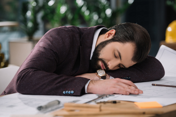 exhausted architect sleeping on building plans at workplace - 写真・画像