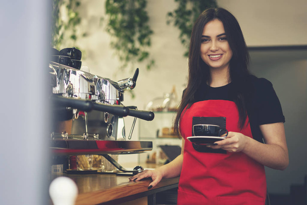 Cheerful waitress enjoying working day in cafeteria making aroma coffee using equipment - 写真・画像