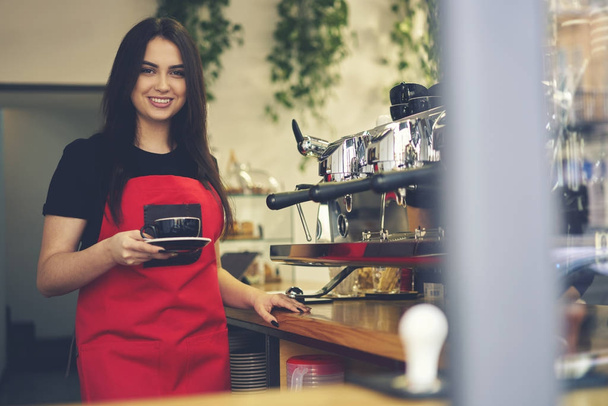 Smiling attractive female waitress enjoying working process holding cappuccino cup - Foto, Bild