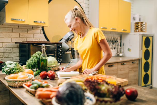 Mladá žena vaření v kuchyni a zápis v knize receptu, zdravé jídlo. Vegetariánská strava, čerstvá zelenina a ovoce na stole  - Fotografie, Obrázek
