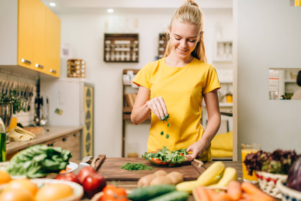 Happy woman cooking salad in kitchen, healthy food. Vegetarian diet, fresh vegetables and fruits on wooden table - Photo, Image