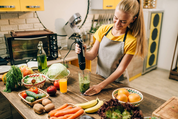 женщина готовит на кухне, смешивая здоровые органические продукты в стекле. Вегетарианская диета, свежие овощи и фрукты на деревянном столе
 - Фото, изображение